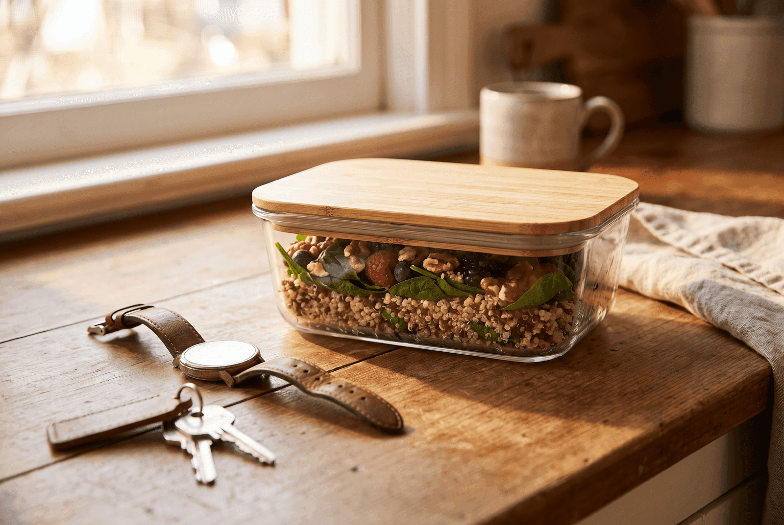 Marmita de vidro pronta com quinoa, folhas verdes e frutas vermelhas, ao lado de chaves e relógio na bancada